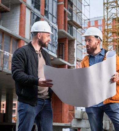 Bearded man in safety helmet holding architectural plan and talking with construction worker outdoors. Male homeowner discussion building plan with specialist at construction site.